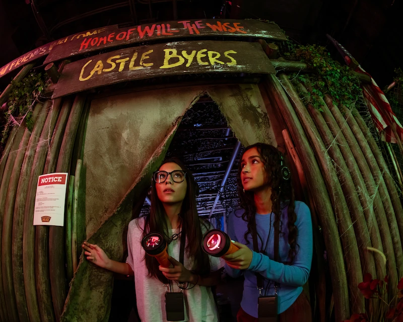 Two friends explore Castle Byers at Netflix House Dallas with headsets and flashlights.