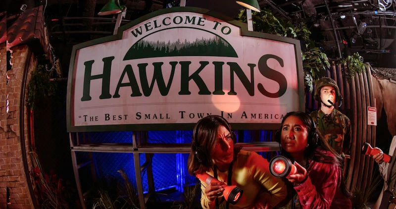 Two friends hold flashlights, wear audio sets, and explore Hawkins.
