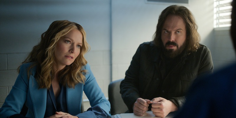 Two people sit at a table in a dimly lit room, appearing serious and focused, possibly in an interrogation or interview setting, with light streaming through blinds creating a tense mood.