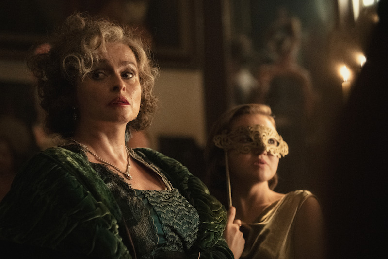 Two women in vintage formal attire at a dimly lit, elegant gathering, one staring intently ahead and the other holding a decorative mask, creating a mysterious and dramatic mood.