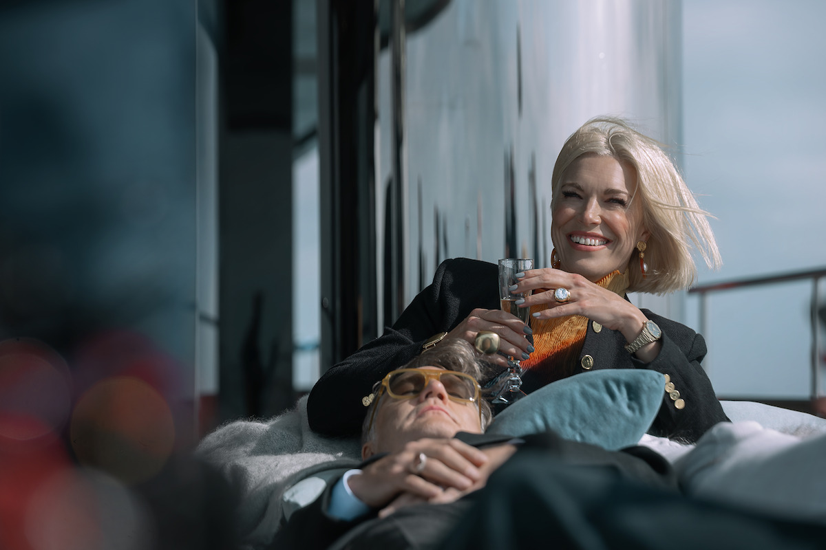 Two people relaxing on a yacht deck, one smiling and holding a drink, the other resting with sunglasses. Bright, luxurious atmosphere with a calm, happy mood, and modern outdoor setting.