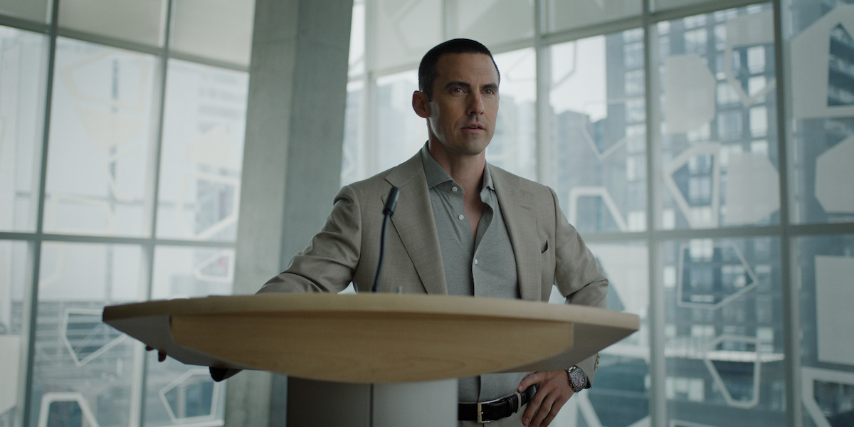 Man in a suit standing at a podium with a microphone in a modern office or conference room, large windows and cityscape in the background.