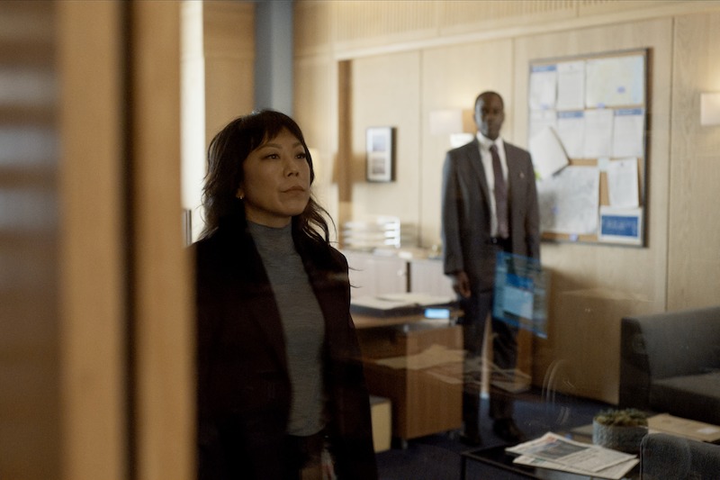 A woman in a blazer stands in the foreground looking serious, while a man in a suit stands in the background of an office with papers pinned to a bulletin board.