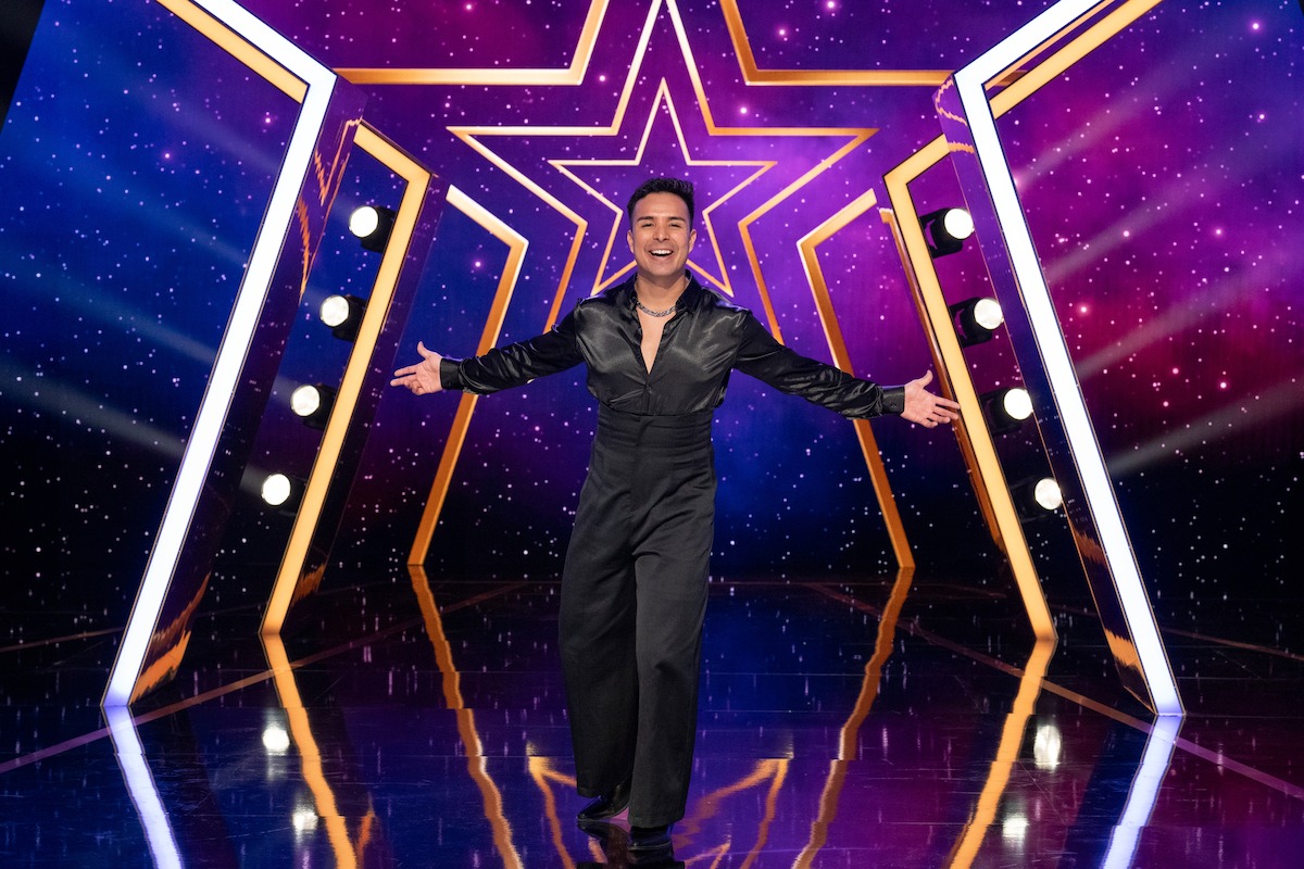 Fernando Velasco in a black outfit stands with arms open on a shiny stage, with a large illuminated star and vibrant purple background with lights.