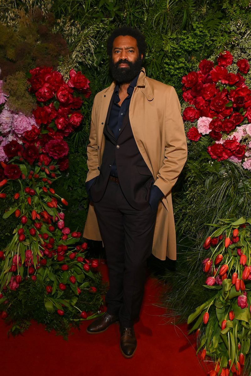 A man in a tan trench coat and dark suit stands on a red carpet surrounded by lush greenery and vibrant red and pink flower arrangements, posing for a photo at a formal indoor event.