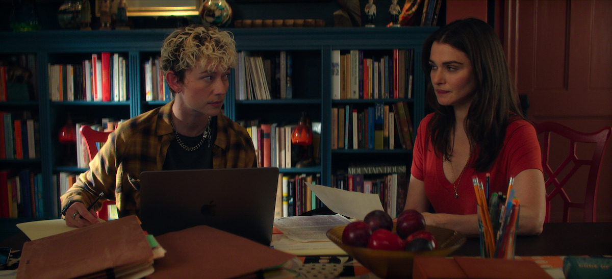 Ellen Robertson as Sid and Rachel Weisz as M sit at a cluttered table with paperwork, folders, and a laptop in a cozy library or study lined with bookshelves, having a focused conversation. A bowl of apples is in the center of the table.