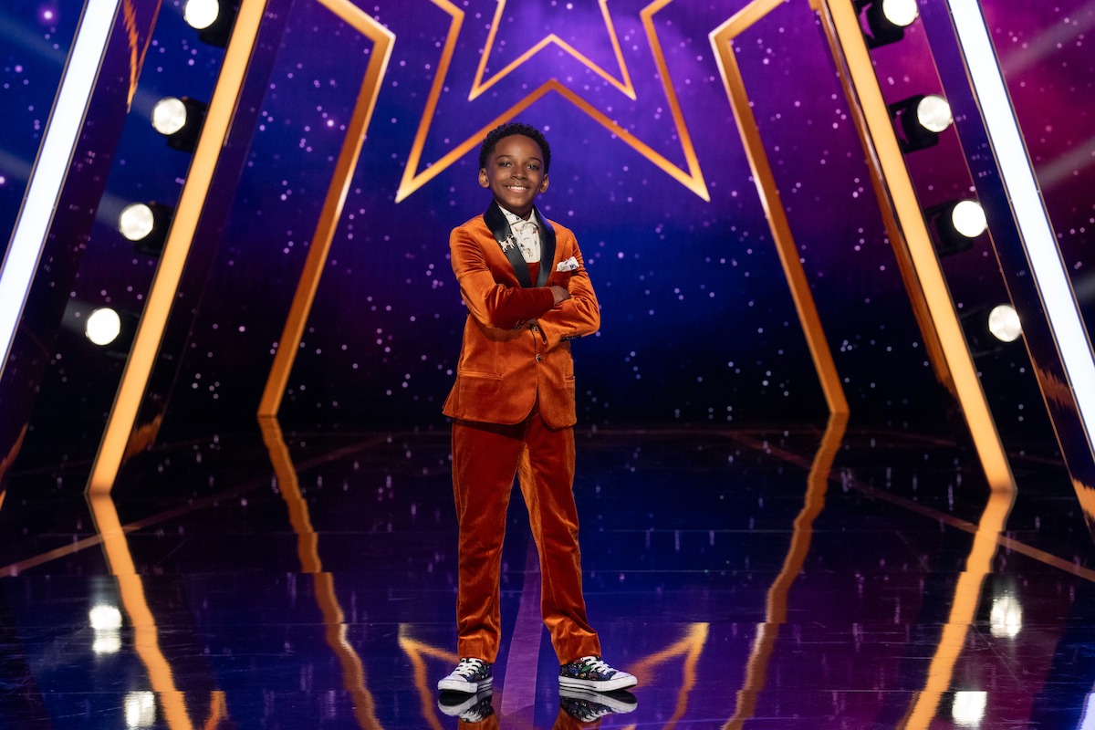 Eric Adrien Williams in a orange suit stands confidently on a shiny stage with a star backdrop and bright lights, suggesting a talent show or performance environment.