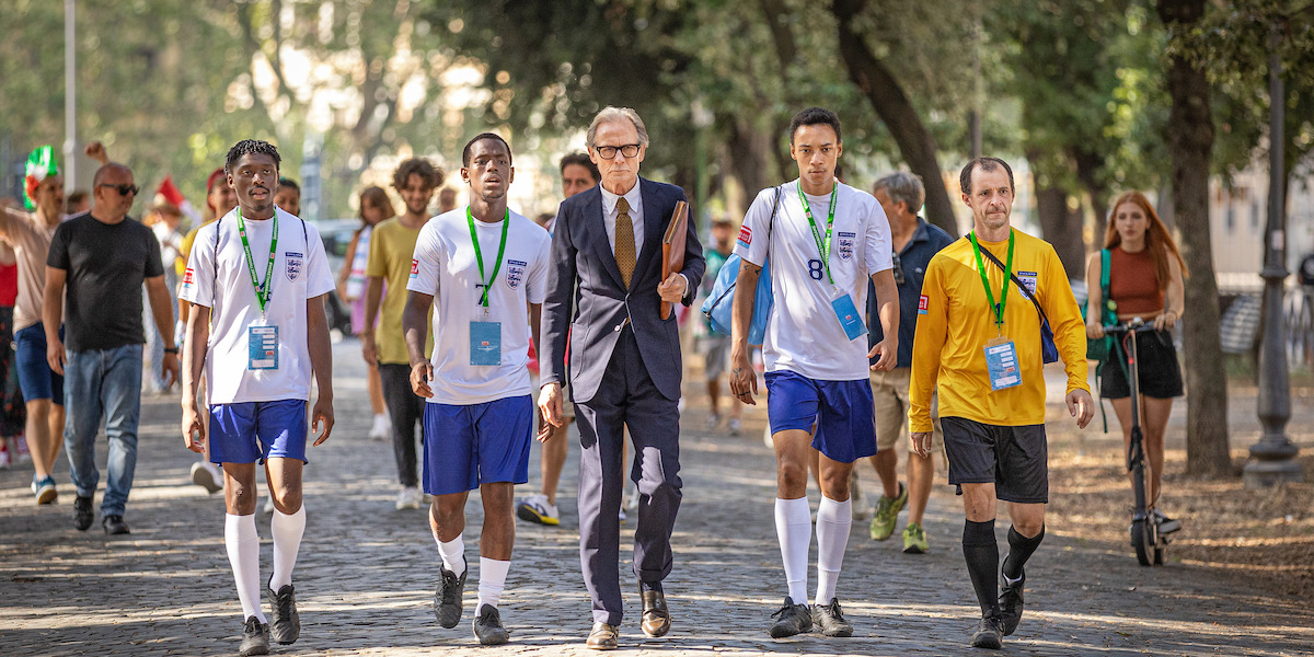 Micheal Ward, Bill Nighy, and the cast of ‘The Beautiful Game’ walk down a street.