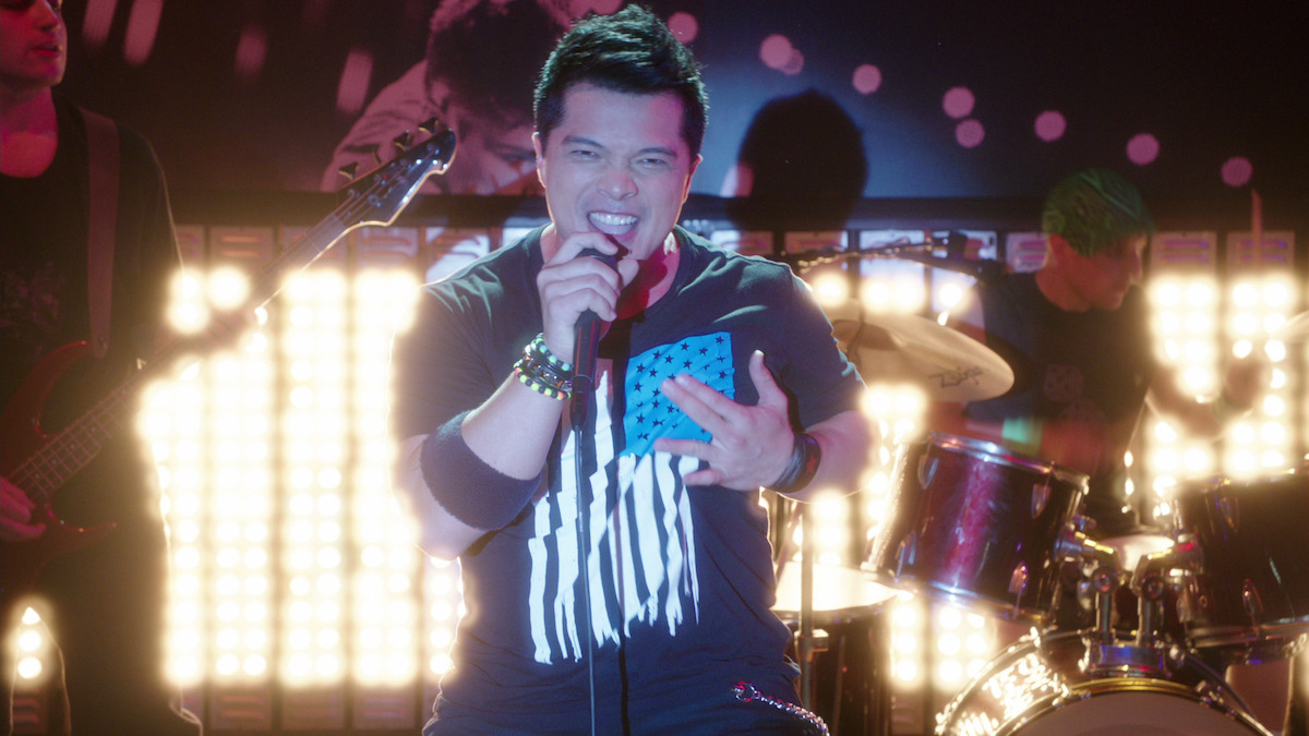 a man in an american flag t-shirt sing with a rock band behind him. 