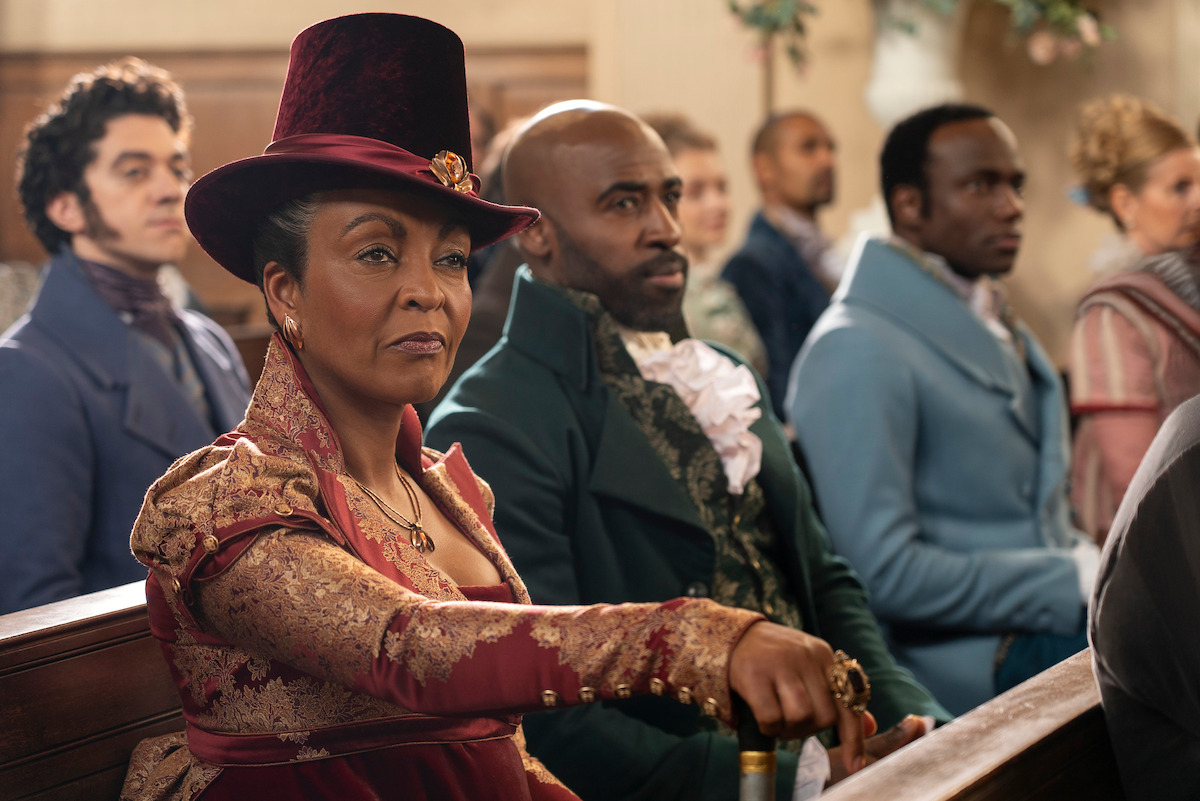 Several people in elegant, historical clothing sit in wooden pews, appearing attentive in a candlelit room, suggesting a formal gathering or ceremony in a period drama setting.