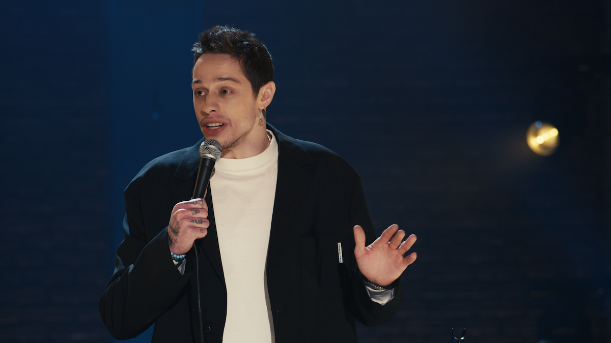 A man performing stand-up comedy on stage, holding a microphone with one hand raised, wearing a dark jacket and white shirt, under stage lighting with a dark blurred background.
