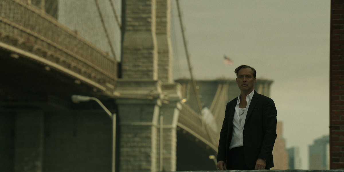 A man in a black suit stands outdoors near a large city bridge with an American flag and city skyline in the background, under cloudy skies. The setting appears urban and subdued.