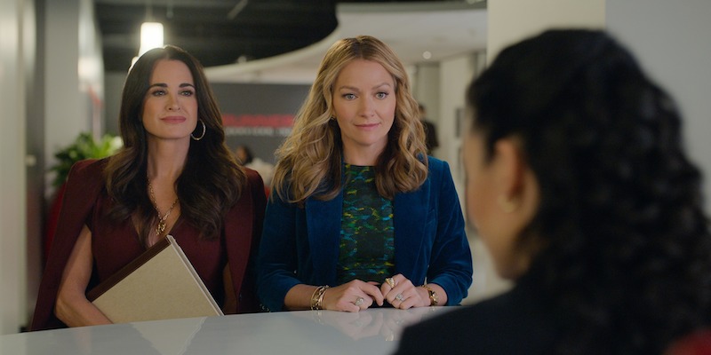 Two professionally dressed women sit across a desk from another person in an office setting, holding folders and appearing engaged in conversation, creating a formal and focused atmosphere.
