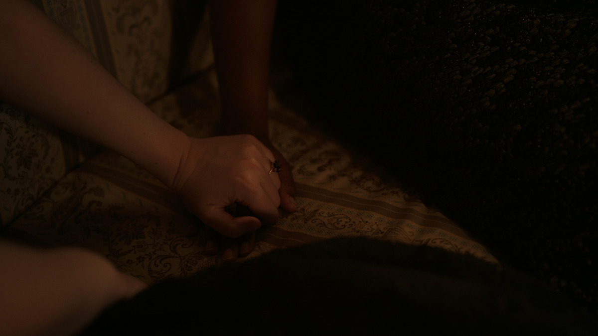 Two hands are intertwined on a patterned fabric surface in a dimly lit, intimate indoor setting.