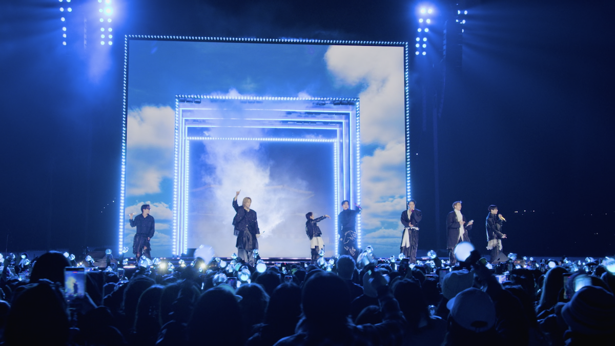 BTS performs on stage with blue lights and a sky-themed digital backdrop, while a large audience with glowing light sticks watches and takes photos in a concert arena.