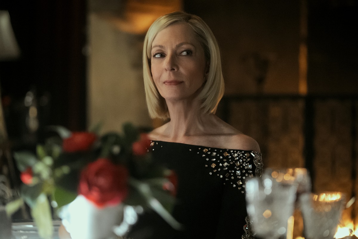 Elegant woman with blond bob haircut in a black, jewel-studded dress sits at a dimly lit, formal dinner table with red roses and crystal glasses, in a luxurious, warm-toned environment.