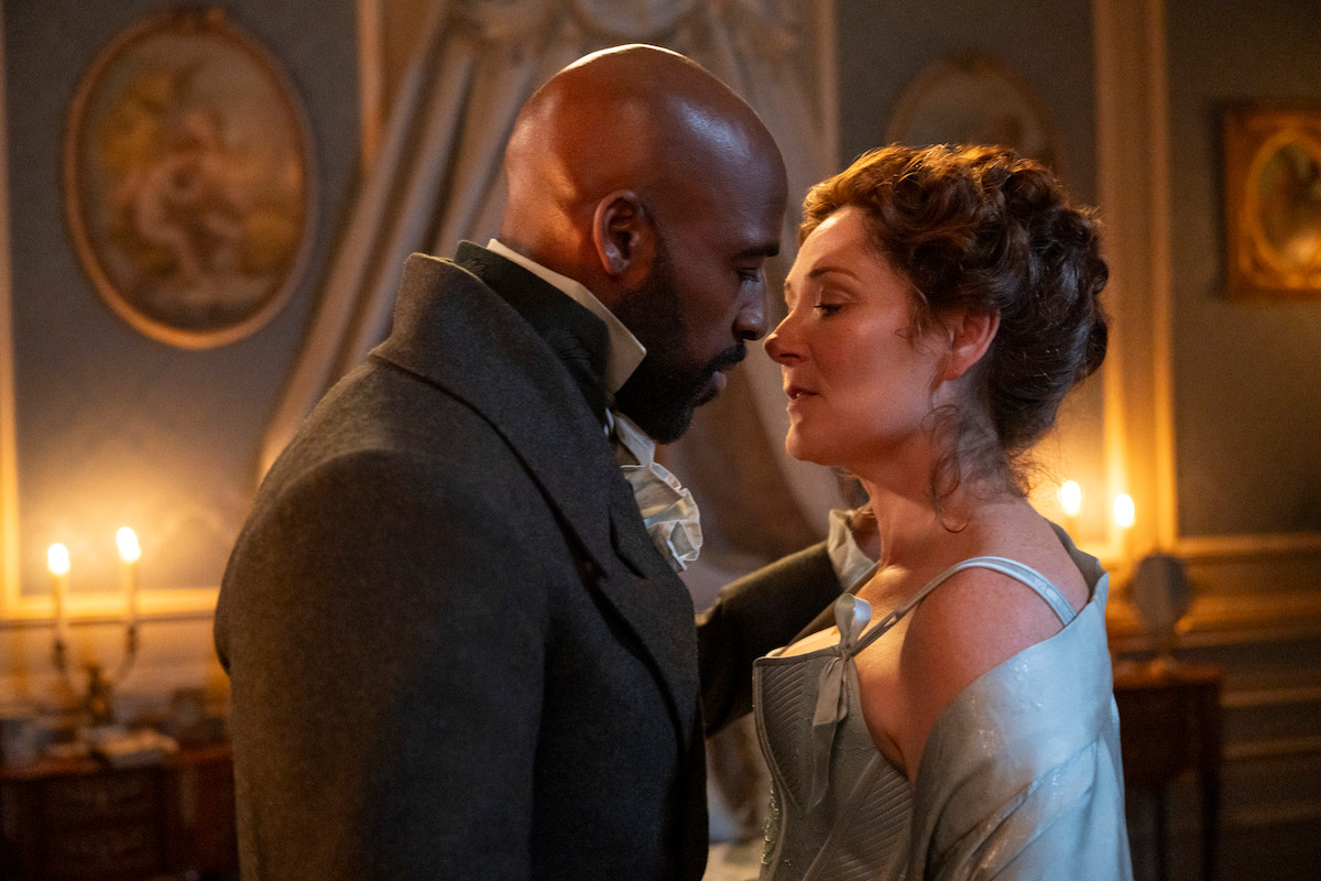 A man and woman in period costumes share an intimate moment in a candlelit, elegant room with ornate walls, framed art, and classic decor, suggesting a romantic or dramatic historical setting.