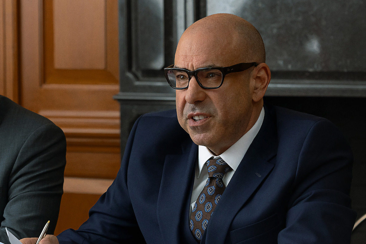 Bald man in glasses and a suit speaking seriously at a meeting in a formal courtroom or office setting, sitting at a table with a pen in hand and wood paneling and a fireplace in the background.