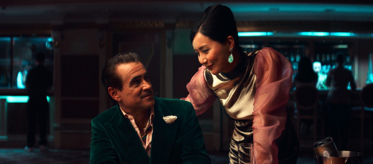 A man in a velvet jacket sits at a casino table while a woman in elegant attire leans in, both engaged in conversation under moody, ambient lighting with a bar in the background.