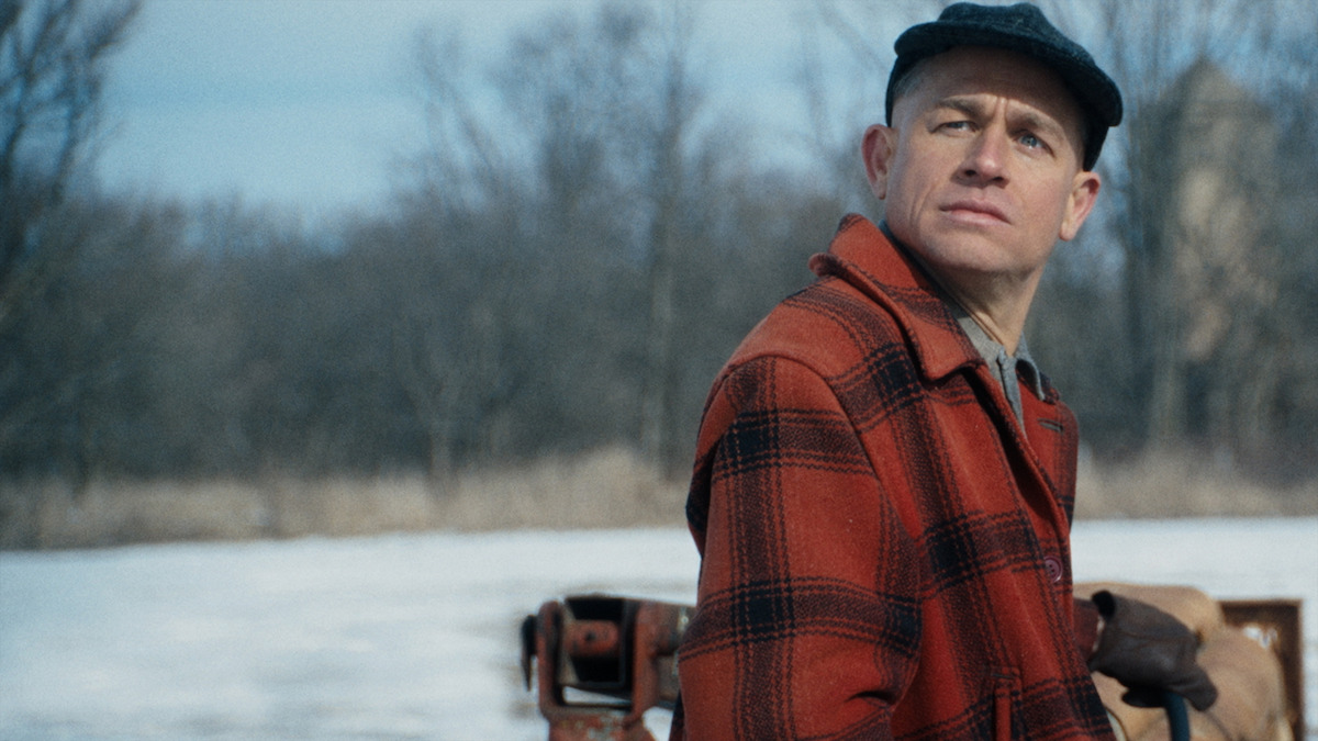 A man in a plaid shirt and baseball cap looks into the distance with a concerned look on his face.