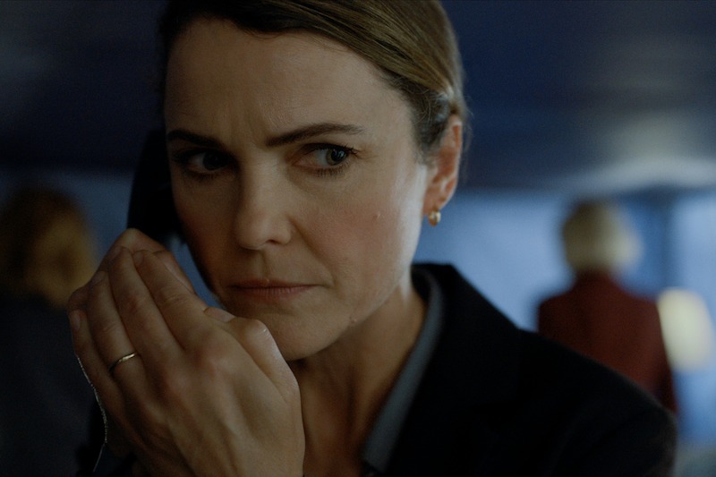 A woman with a serious expression holds a phone to her ear and covers her mouth with her hand, appearing to listen intently in a dimly lit room.