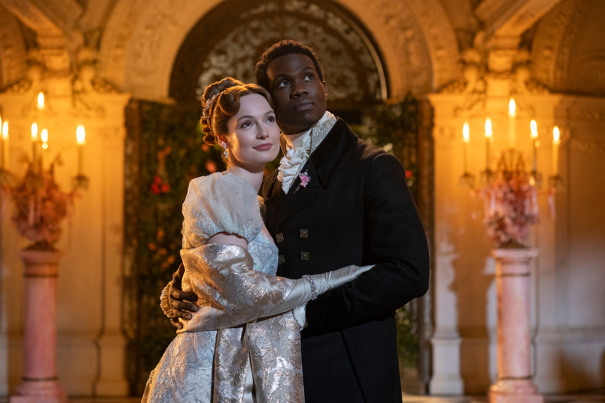 A couple in elegant period costumes stand embraced in a grand, ornately decorated hall with warm candlelight and floral arrangements, suggesting a romantic or formal event in a historical setting.