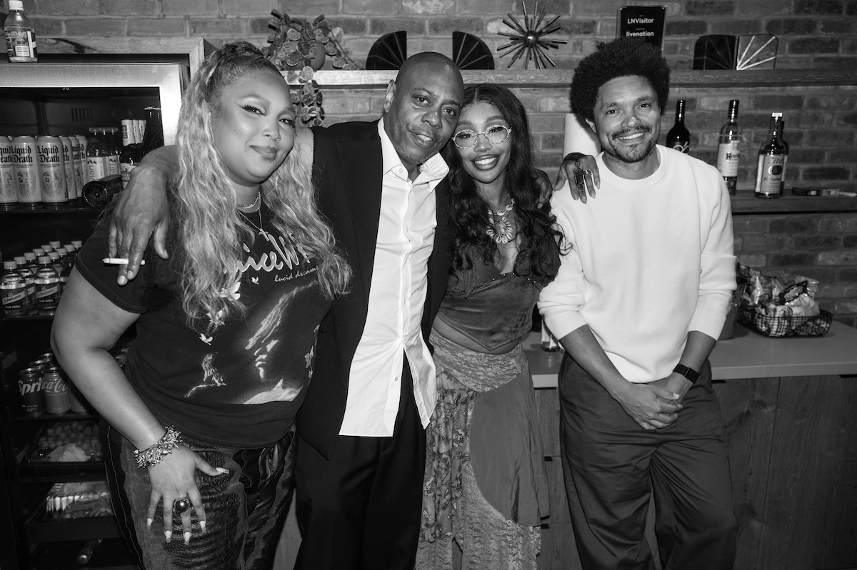 Four people pose together, smiling in a casual indoor setting by a brick wall, with drinks and snacks on shelves behind them, giving a relaxed and celebratory atmosphere.
