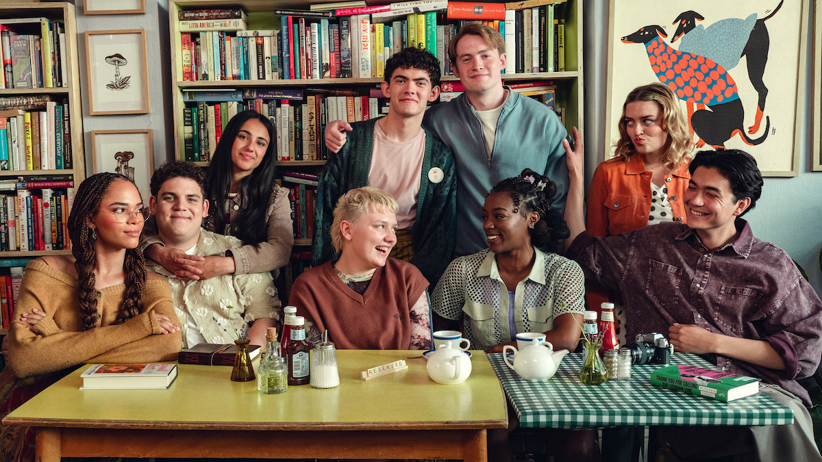 A group of eight young adults sitting and standing around tables with tea and books in a cozy room filled with bookshelves and colorful art, smiling and enjoying each other's company.