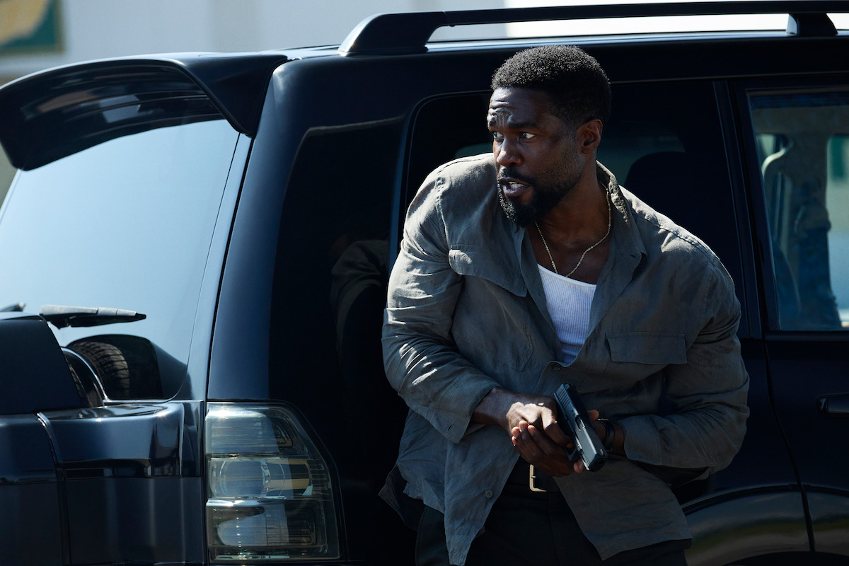 Man cautiously exits black SUV holding a handgun, wearing a gray jacket and white shirt, appearing alert in a tense urban outdoor setting.