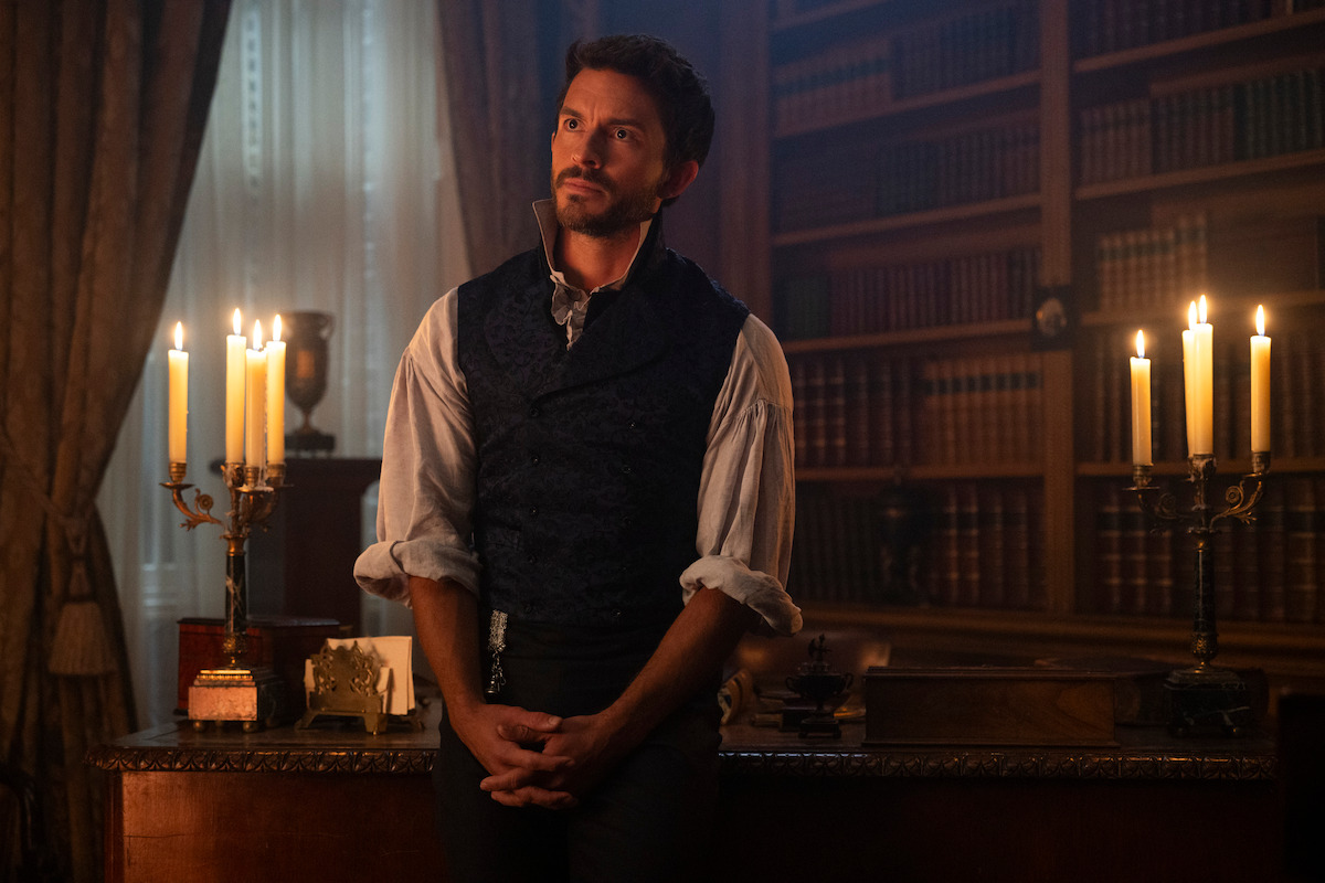 A man in historical clothing leans against a wooden desk in a richly decorated library with shelves of books, lit by several candelabras. The ambiance is warm and atmospheric with vintage decor and heavy drapes in the background.