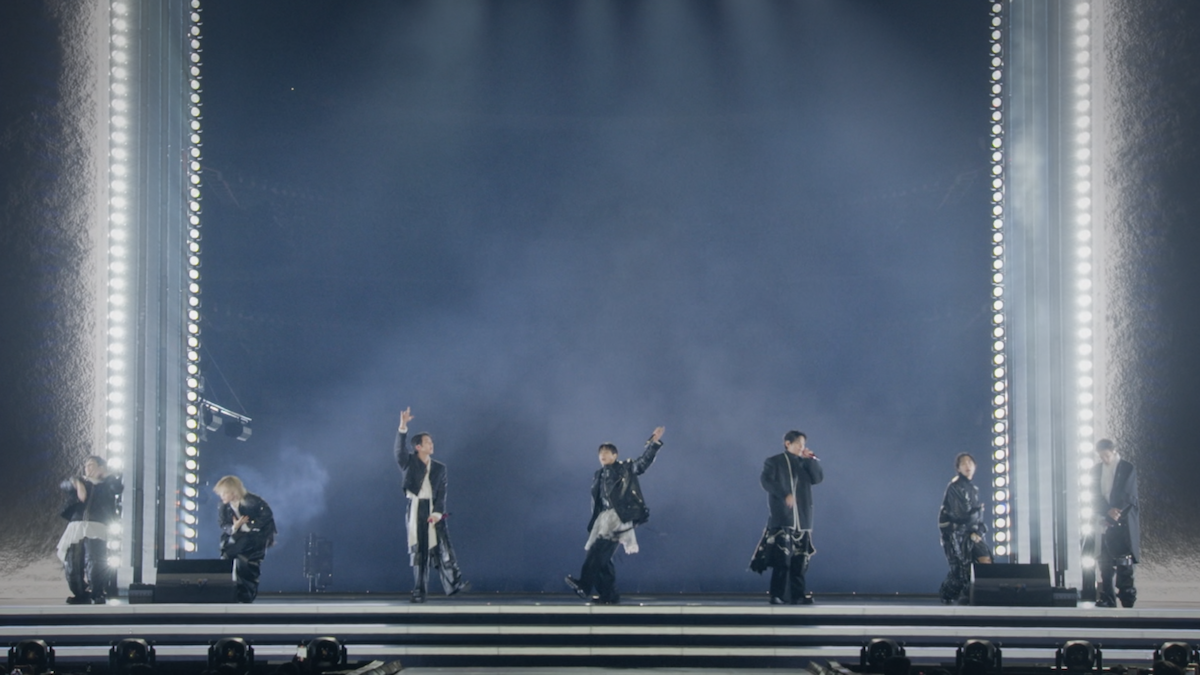 Seven performers on stage with dramatic lighting, vertical light columns, and a smoke-filled background, singing and gesturing energetically in a large concert venue.
