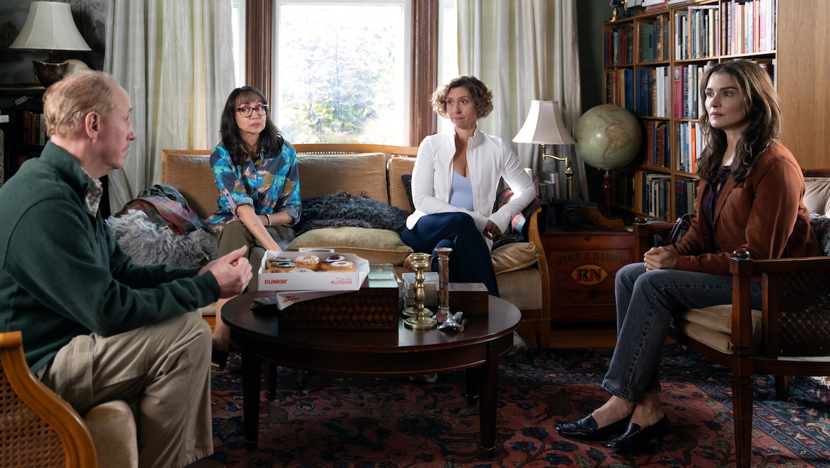 Four people sit in a cozy living room with bookshelves, a globe, large windows, and a coffee table with snacks, appearing to have a serious conversation.