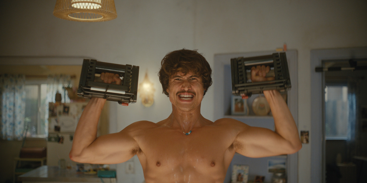 Shirtless man lifting adjustable dumbbells in a home kitchen, sweating and smiling with determination, surrounded by warm lighting and domestic decor.