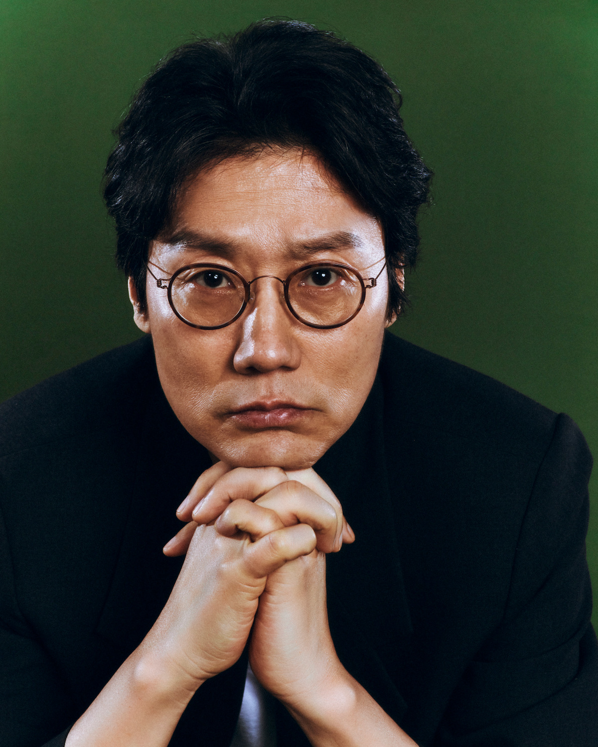 A man with glasses and dark hair sits against a green background, wearing a dark jacket. He looks serious, resting his chin on his hands, gazing directly at the camera in a studio portrait setting.