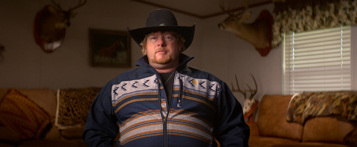 A man in a cowboy hat and patterned jacket sits in a living room with hunting trophies and mounted animal heads on the walls, brown couches, and decorative pillows, lit by natural light from a window with patterned curtains.