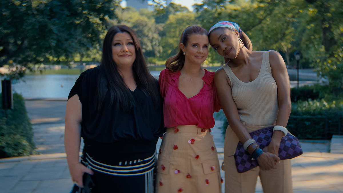 Three women standing together outdoors in a park, posing for a photo on a sunny day with trees and a pond in the background.