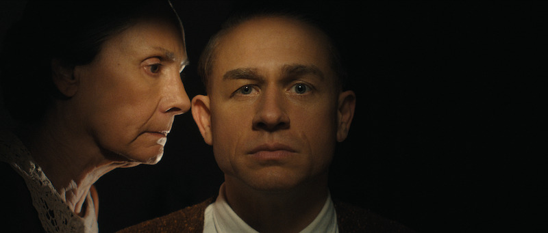 An older woman leans in closely to whisper into the ear of a serious-looking man who stares straight ahead in a dark setting.