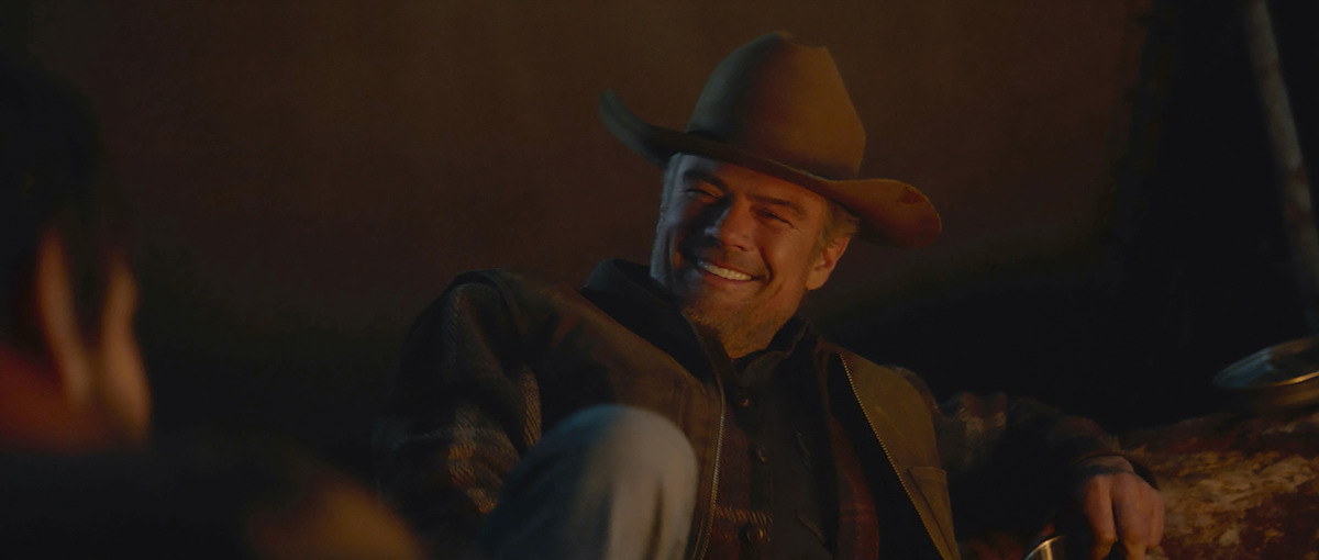 A man in a cowboy hat and jacket smiles while sitting by a campfire at night, appearing relaxed outdoors. Warm firelight lights his face, and another person is partially visible in the foreground.