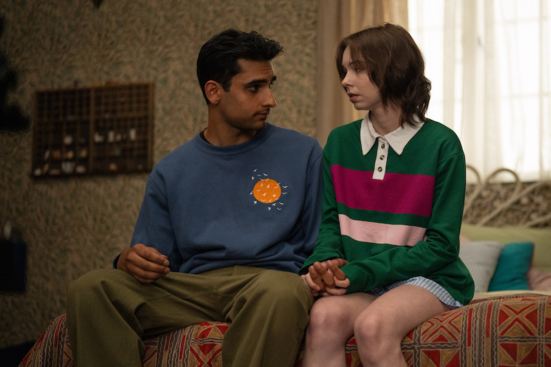 Two people sit closely together on a bed in a cozy, softly lit bedroom, looking at each other and holding hands, suggesting a serious or intimate conversation. The room has patterned wallpaper and soft furnishings.