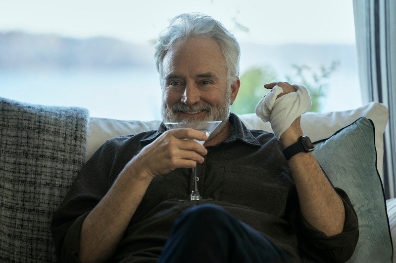 A smiling older man with gray hair and a beard sits on a couch holding a martini glass in one hand and a cloth in the other, looking relaxed and content.