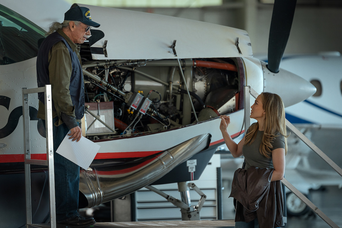 A man and a woman discuss beside an aircraft with its engine exposed inside a hangar, suggesting a maintenance or technical check setting.