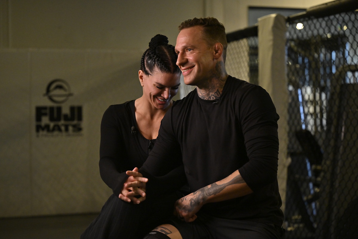 Two people sitting and smiling together inside an MMA gym, with a FUJI MATS logo on the wall and part of an octagon cage visible in the background, both dressed in black athletic wear.