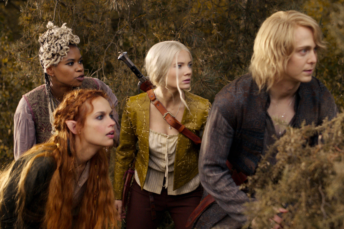 Four fantasy characters in medieval-style clothing crouch together in a dense, wooded forest, looking alert and cautious as if on a quest or facing danger, creating a tense and adventurous atmosphere.