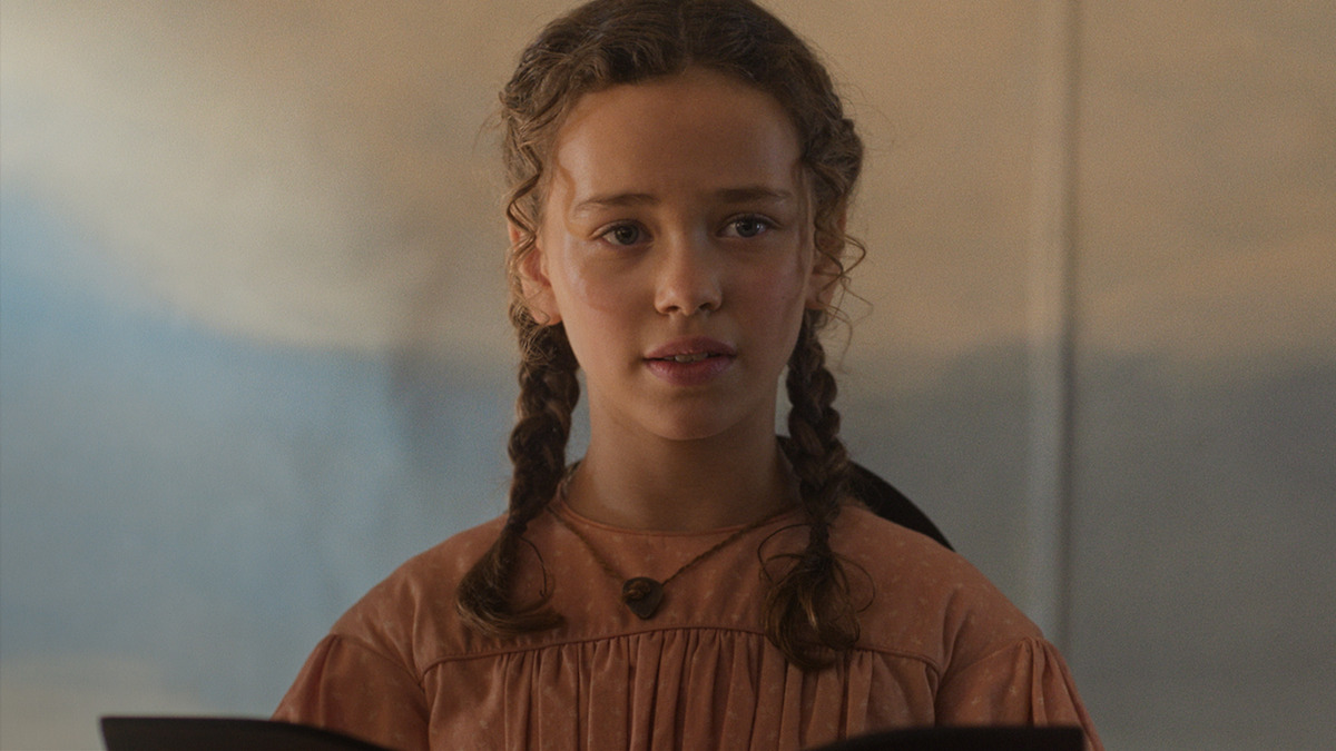 Young girl with braided hair wearing a brown dress stands indoors, holding papers and looking forward, with a soft-focus background suggesting a calm, warm environment.