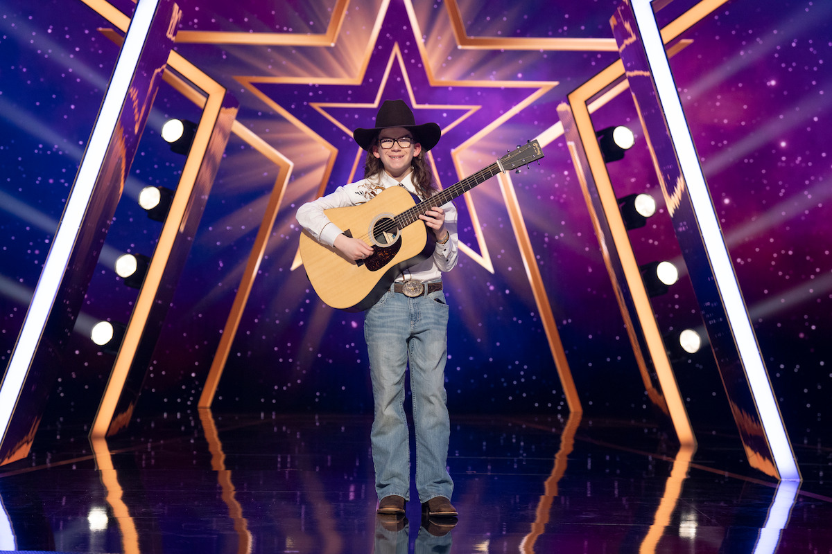 Myles Gee in cowboy hat and boots playing acoustic guitar on a brightly lit stage with star decorations and a purple background, likely during a talent competition.