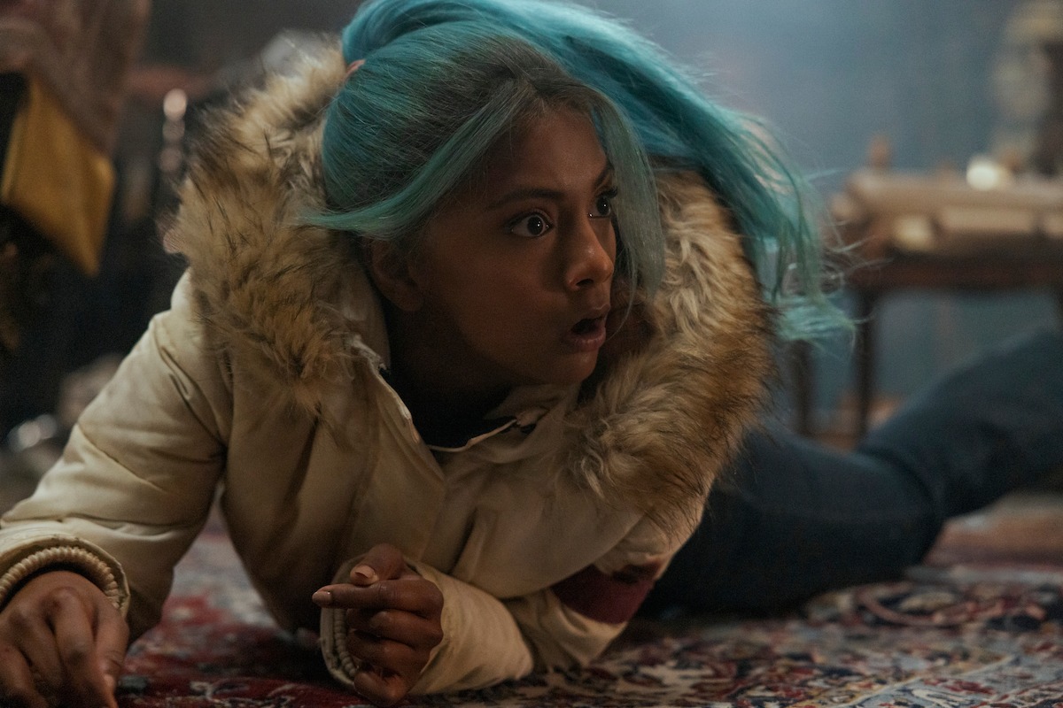 A surprised young woman with turquoise hair in a white fur-lined jacket is lying on a patterned rug in a dimly lit room, appearing alarmed or cautious.