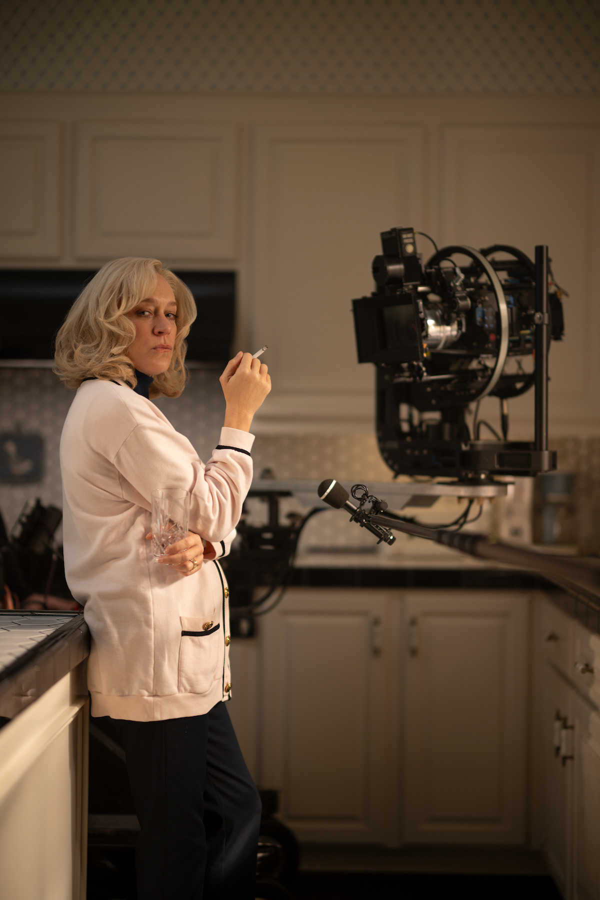 Behind the scenes with Chloë Sevigny as Kitty Menendez. She wears a white top and stands in her kitchen smoking a cigarette.