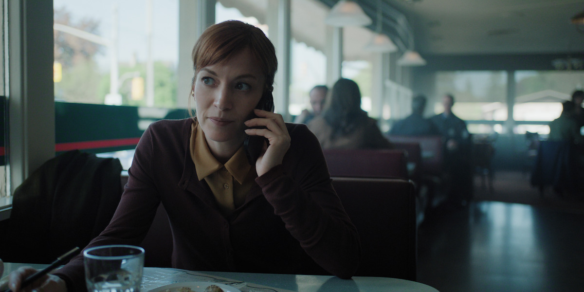 A woman with red hair sits in a diner booth talking on her phone, with a glass of water and a plate of food on the table; the diner is softly lit with other patrons in the background.