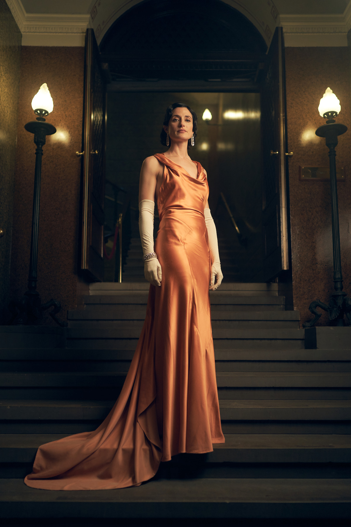 Lizzie Shelby stands on a suitcase in a sleek coral amber silk dress and gloves in 'Peaky Blinders'
