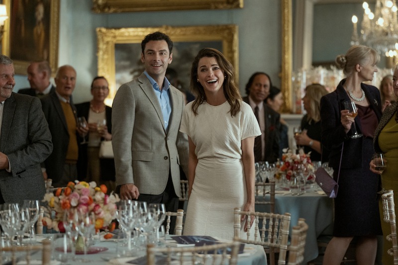 A man in a light suit and a woman in a white dress stand smiling and looking ahead at a formal event, surrounded by guests holding drinks in an elegantly decorated room.